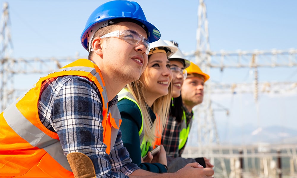 Porträtgruppe junger Menschen mit Schutzhelmen schauen in die Ferne vor elektrischen Komponenten eines Stromerzeugungskraftwerks